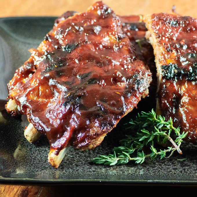 Grilled Baby Back Ribs covered with BBQ sauce on a black plate with a sprig of thyme 
