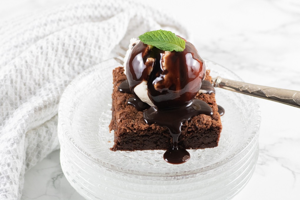 Chocolate brownie square on a stack of glass plates with a fork, a scoop of ice cream chocolate sauce and a mint leaf
