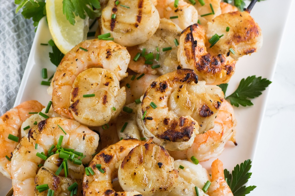 Grilled scallops and shrimp on a white plate with chopped chives, parsley and lemon wedge