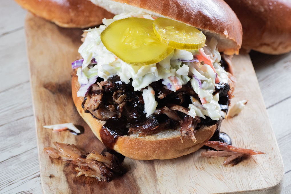 Smoked Pulled pork sandwich on a wooden board with coleslaw, BBQ sauce, and pickles