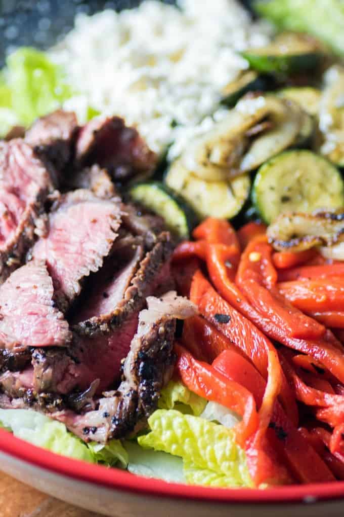Sliced grilled steak with lettuce, roasted red peppers, and grilled zucchini in a red and white bowl
