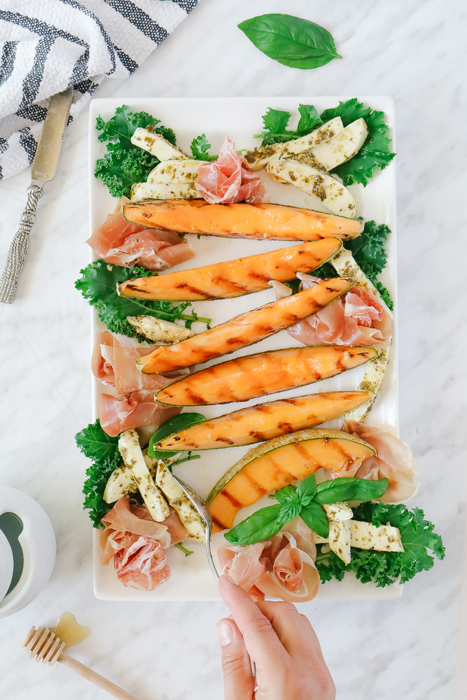 Grilled cantaloupe slices on a white platter with prosciutto and mozzerella slices