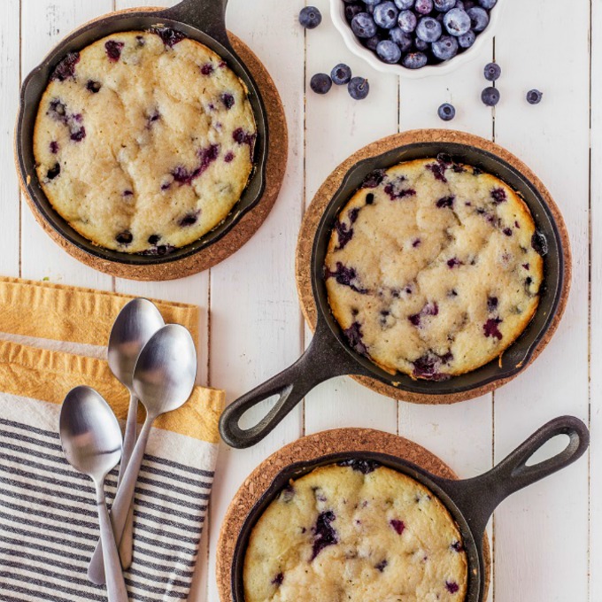 Lemon Blueberry buttermilk cake in small cast iron skillets on a wooden board with spoons and a bowl of blueberries.