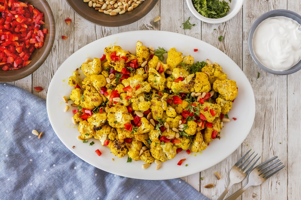 White platter with curry grilled cauliflower garnshed with diced red peppers, toasted pine nuts, and parsley