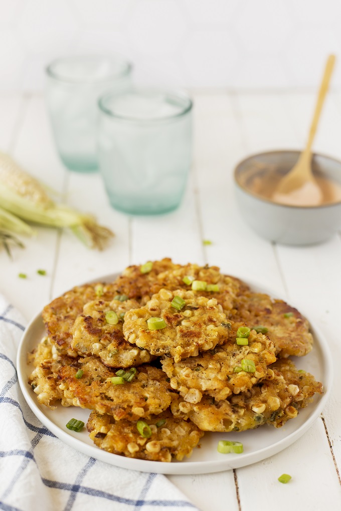 Stack of cooked corn fritters on a white plate garnished with sliced green onion