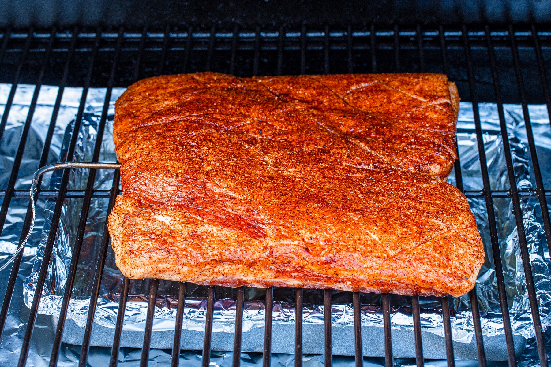Whole Pork belly covered with dry rub just placed onto the smoker grill with a temperature probe inserted into the meat.