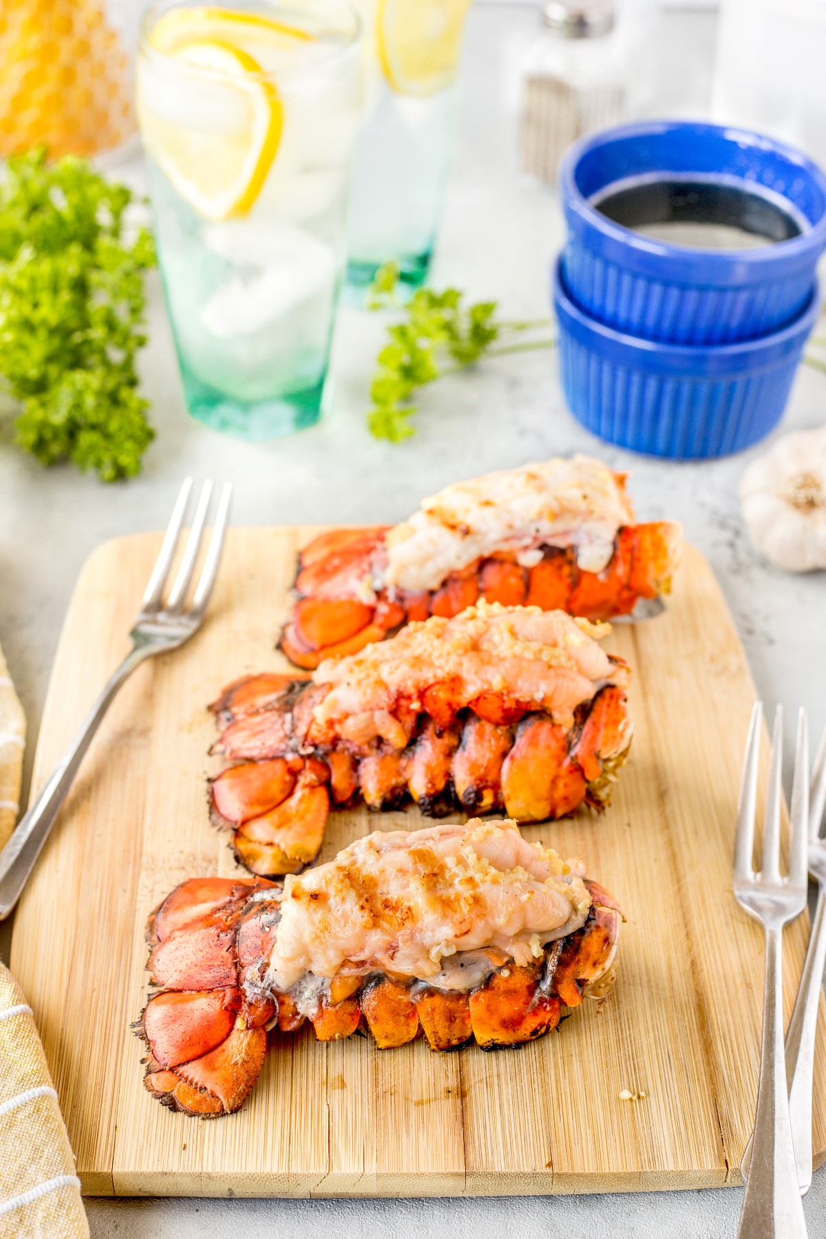 3 grilled lobster tails on a wooden cutting board with 3 forks laying beside them.
