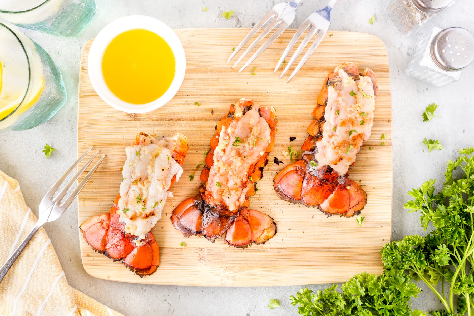 Grilled lobster tails on a wooden cutting board with a small bowl of melted butter and 3 forks, a bunch of parsley beside the board.