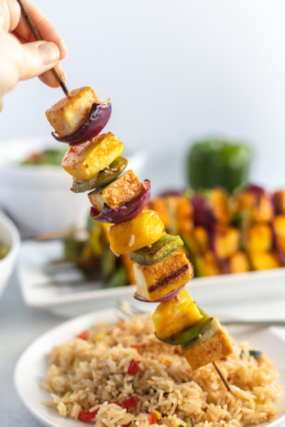 Cooked Hawaiian grilled tofu skewer being held up over a plate of fried rice
