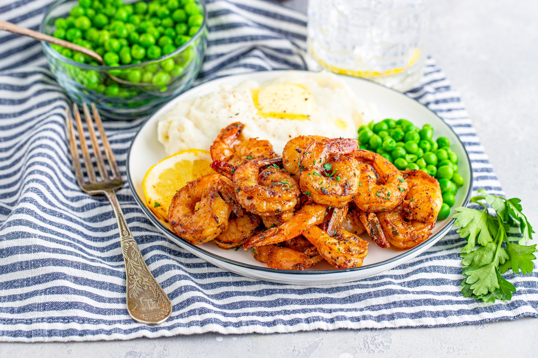 Blackened shrimp on a white dinner plate with peas, and mashed potatoes on a blue and white striped place mat.