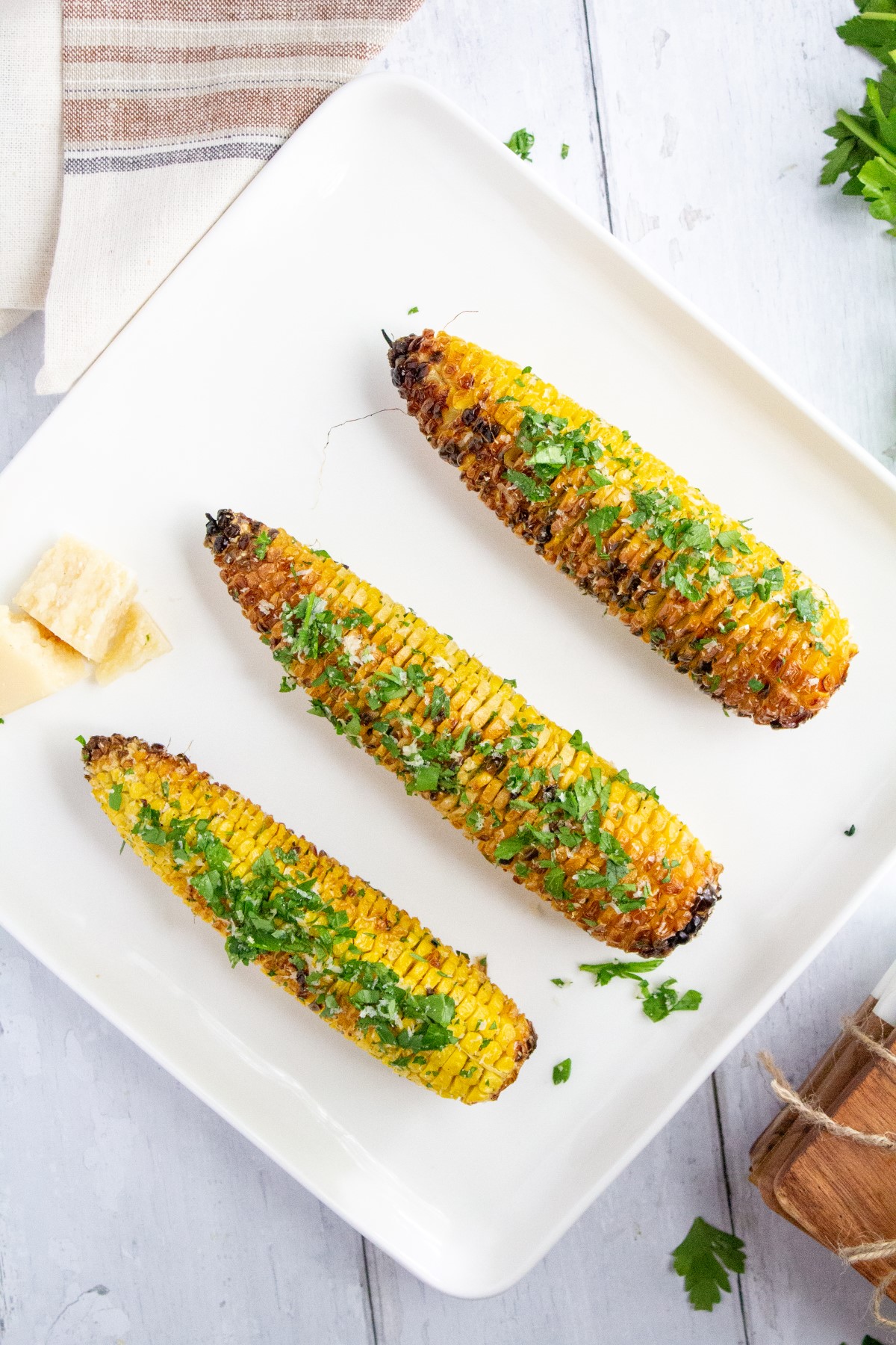 3 grilled cobs of corn on a white platter covered in grated parmesan, chopped parsley and minced garlic.