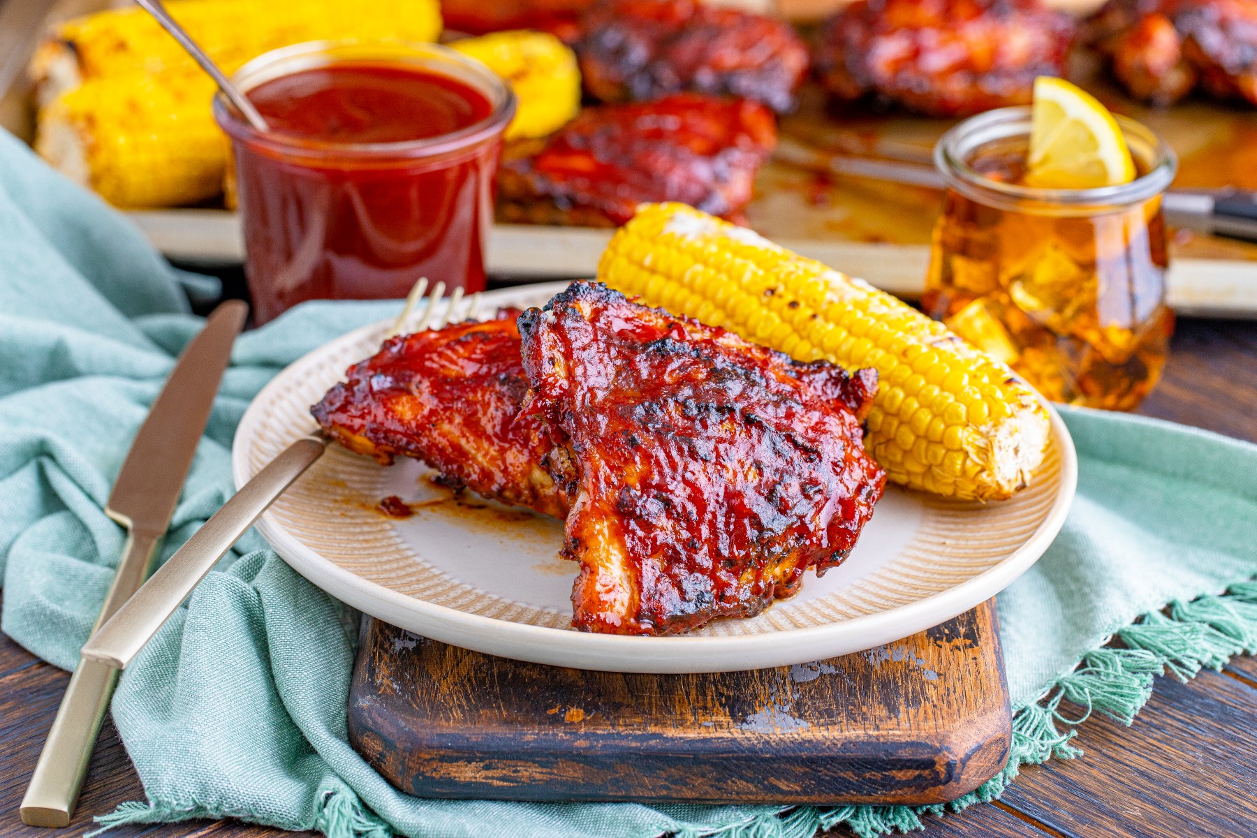 Grilled BBQ chicken thighs covered in BBQ sauce on a white plate with a grilled cob of corn beside the chicken thighs