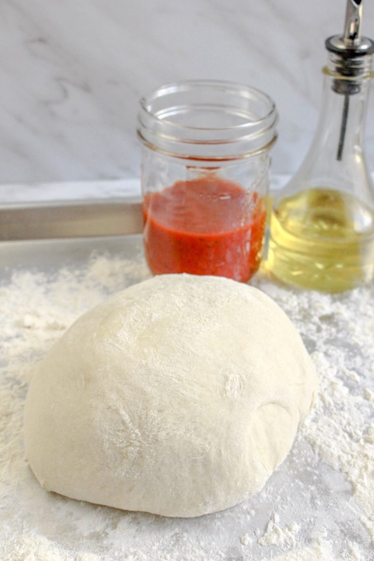 Ball of pizza dough on a floured surface with a jar of pizza sauce and a bottle of oil behind