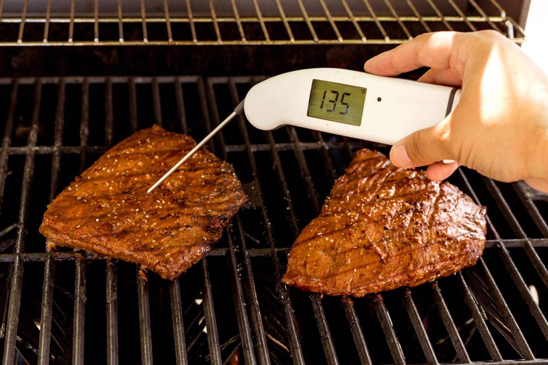 Marinated flank steak cooking on the grill, with a instant read thermometer that reads 135 degrees F.