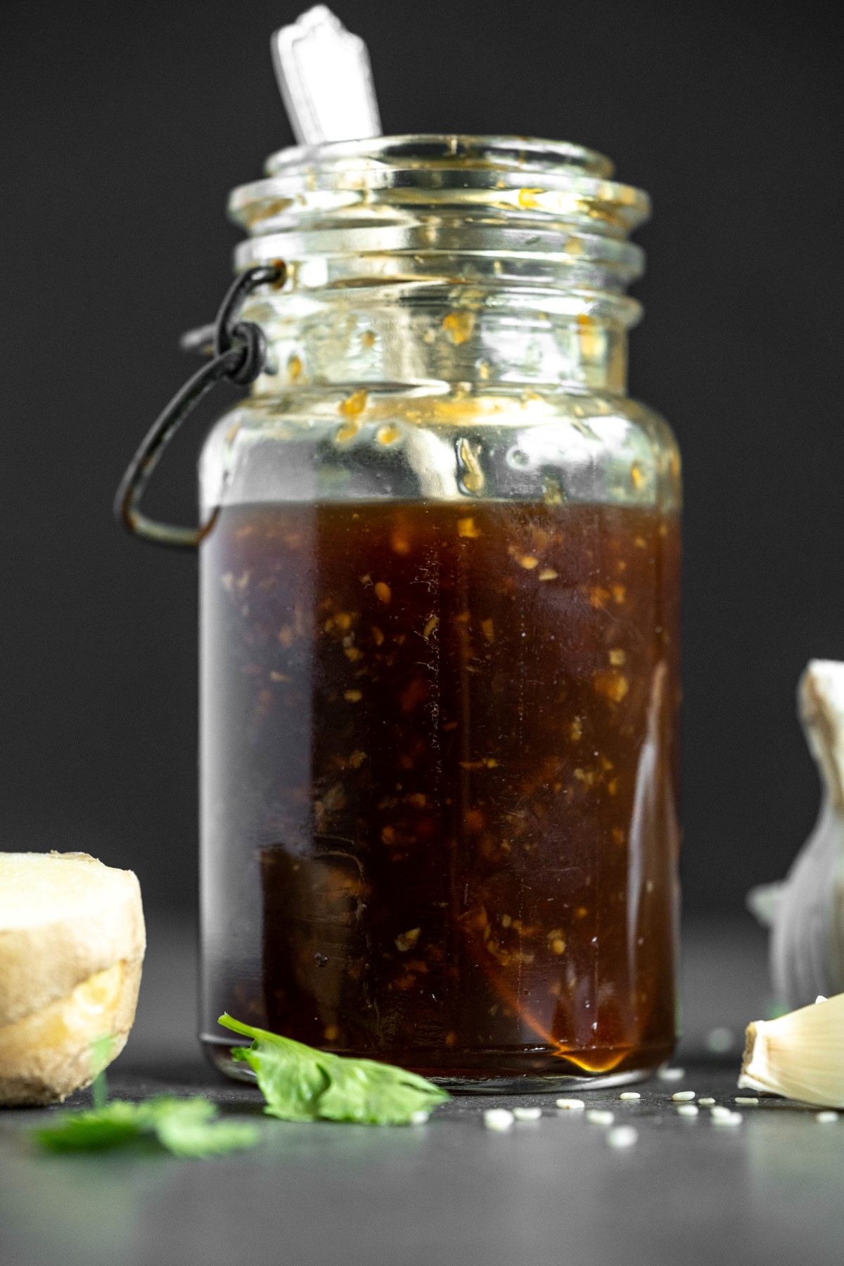 Teriyaki sauce in a clear glass jar with sesame seeds, a piece of ginger and clove of garlic beside the jar.