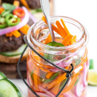 Quick pickled vegetables in a glass jar with a fork in the jar and some burgers topped with the pickled veggies in the background