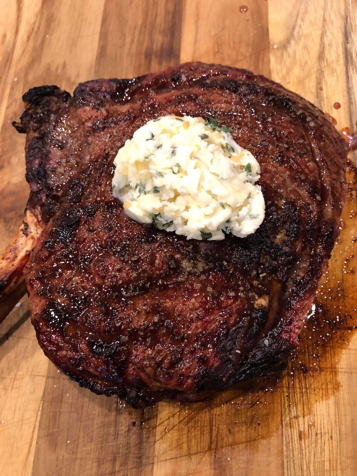 Grilled tomahawk steak resting on a wooden cutting board with a large dollop of garlic herb butter on top.