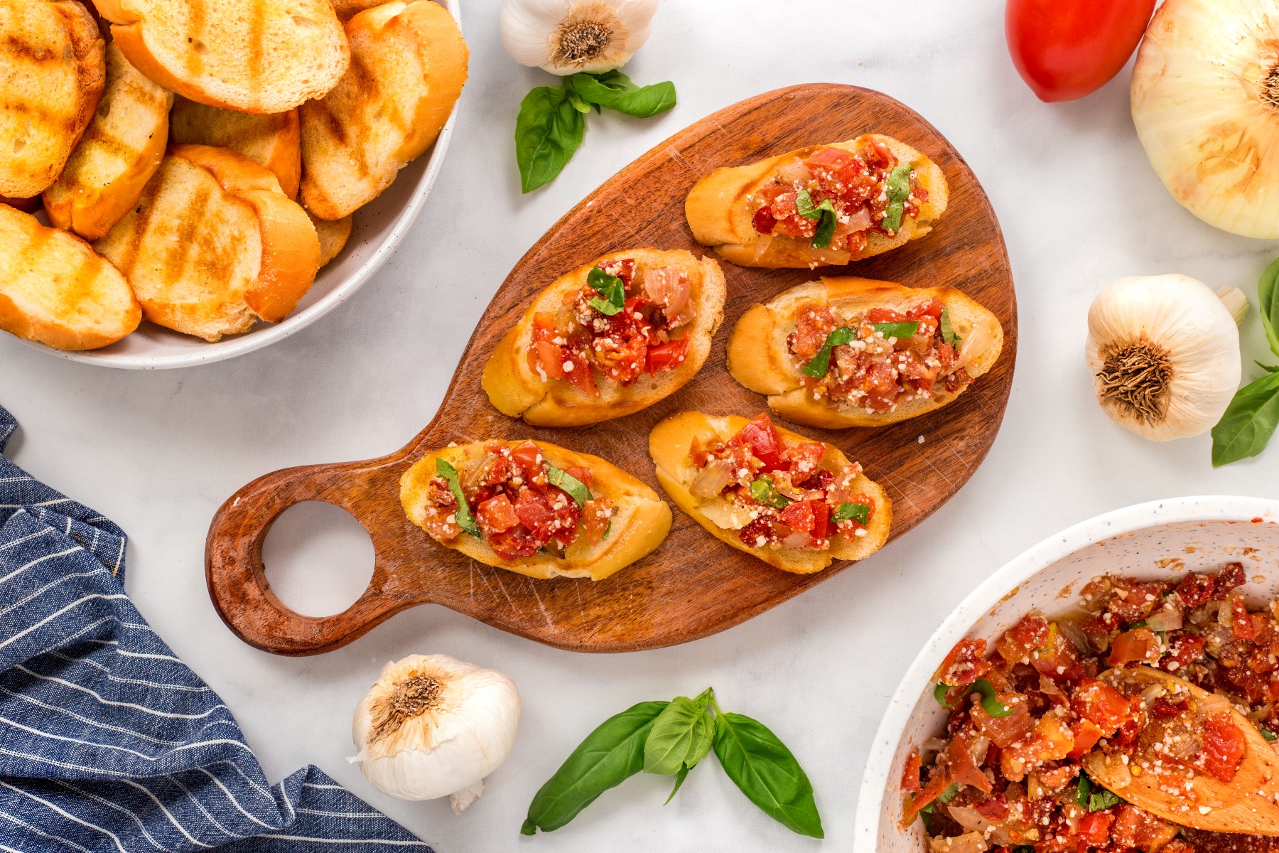 Grilled tomato bruschetta served on a wooden board with extra toasts and a bowl of bruschetta beside the board.