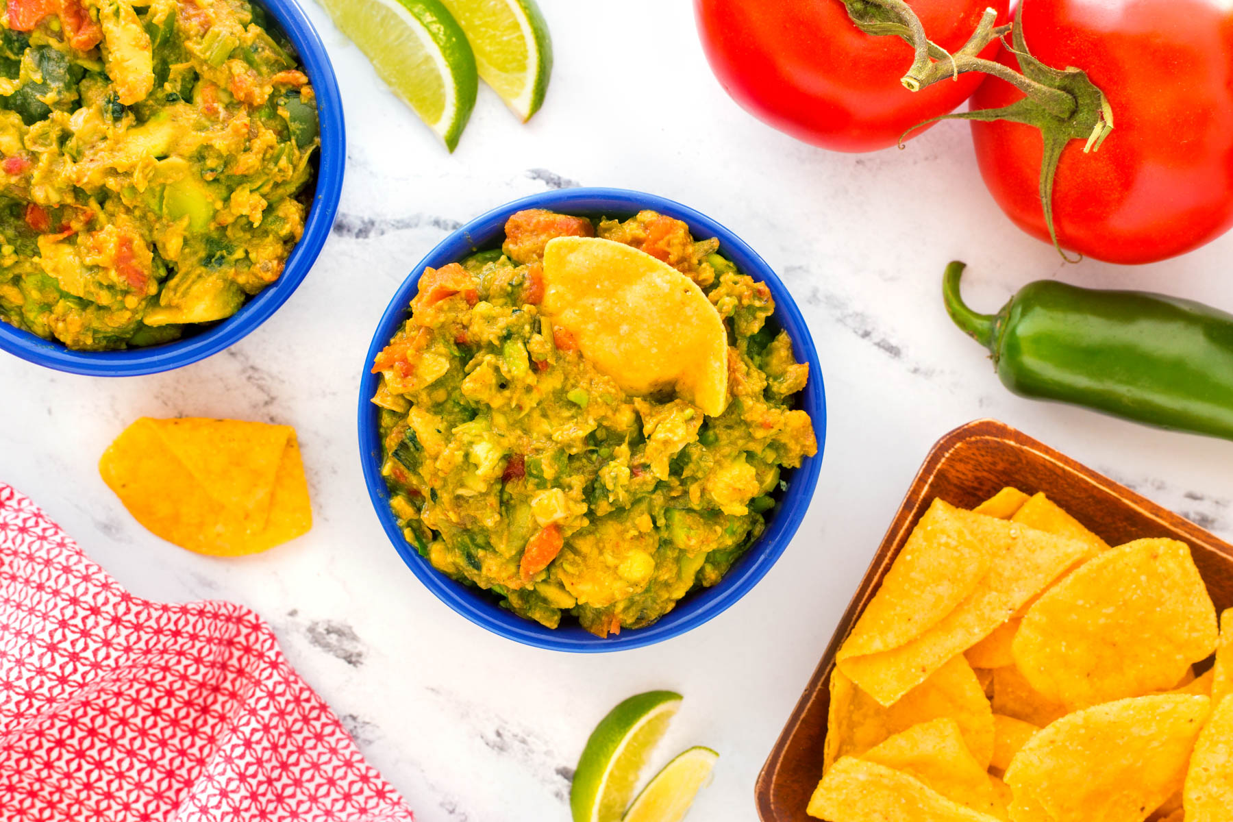 Grilled guacamole in a small blue serving bowl with tortilla chips, lime wedges, whole jalapenos and tomatoes around the bowl