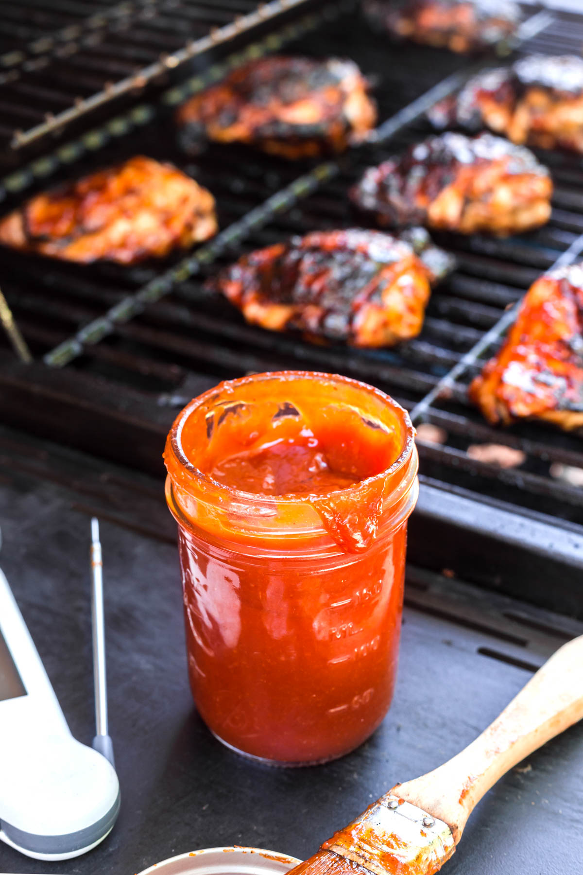 Peach bourbon BBQ sauce ins glass jar beside the grill that is cooking chicken thighs.