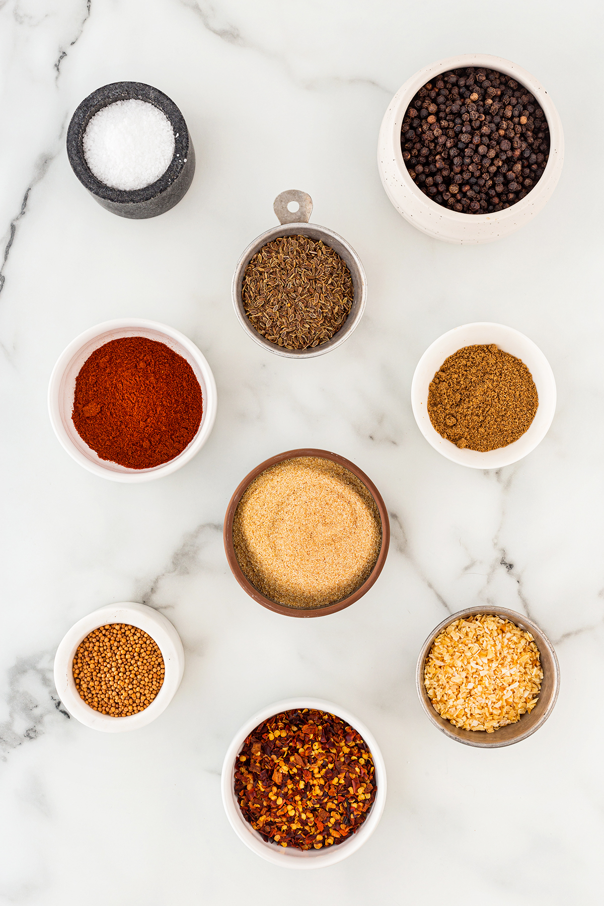 All the ingredients to make Montreal Steak Seasoning in small bowls on a marble countertop.