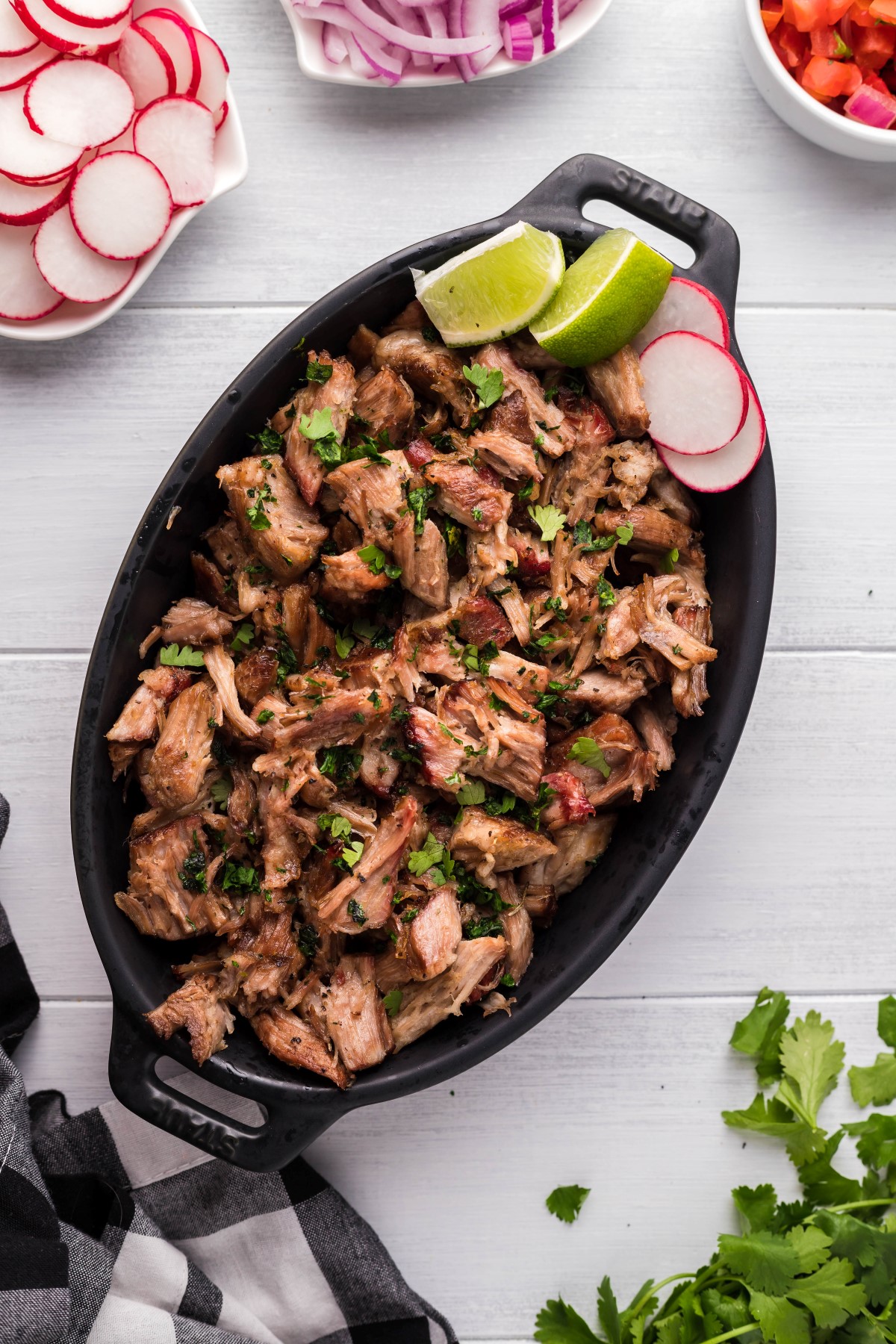 Slow smoked pork shoulder pulled and placed in an oval black serving dish with chopped parsley on top and sliced radishes and lime wedges. 