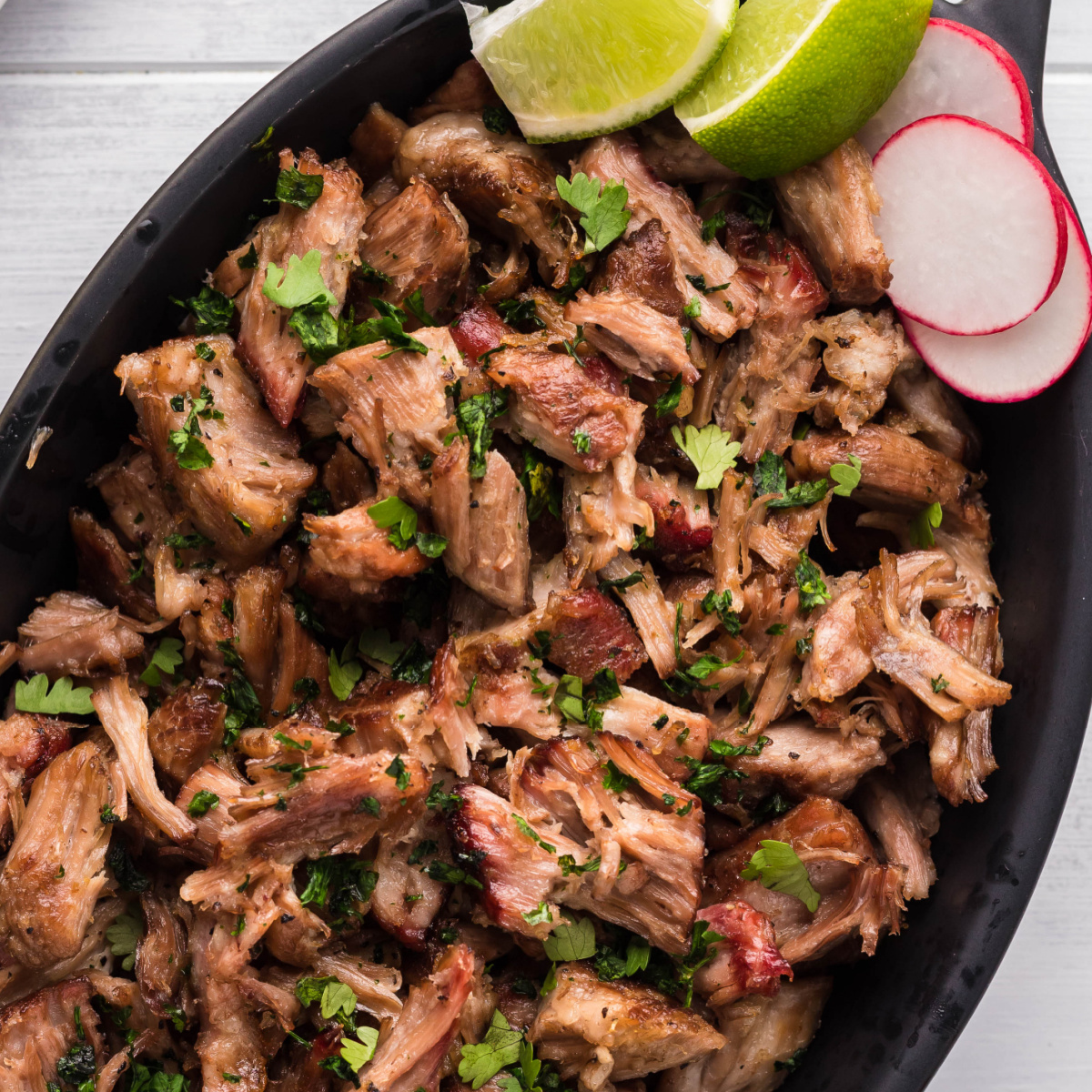 Smoked carnitas in an oval dish with slices of radish and lime wedges, garnished with chopped parsley.