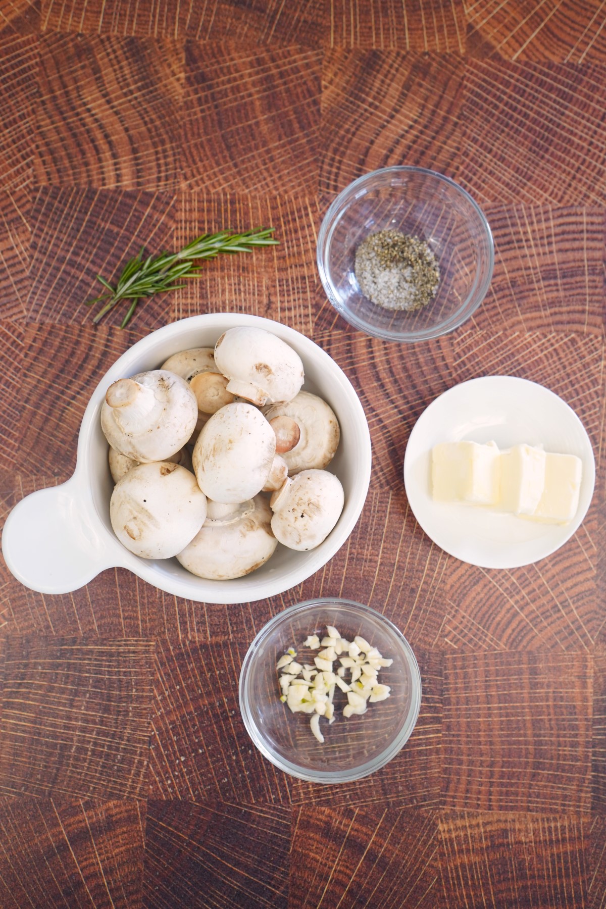 Ingredients to make Rosemary Smoked Mushrooms in small glass bowls on a wooden cutting board.