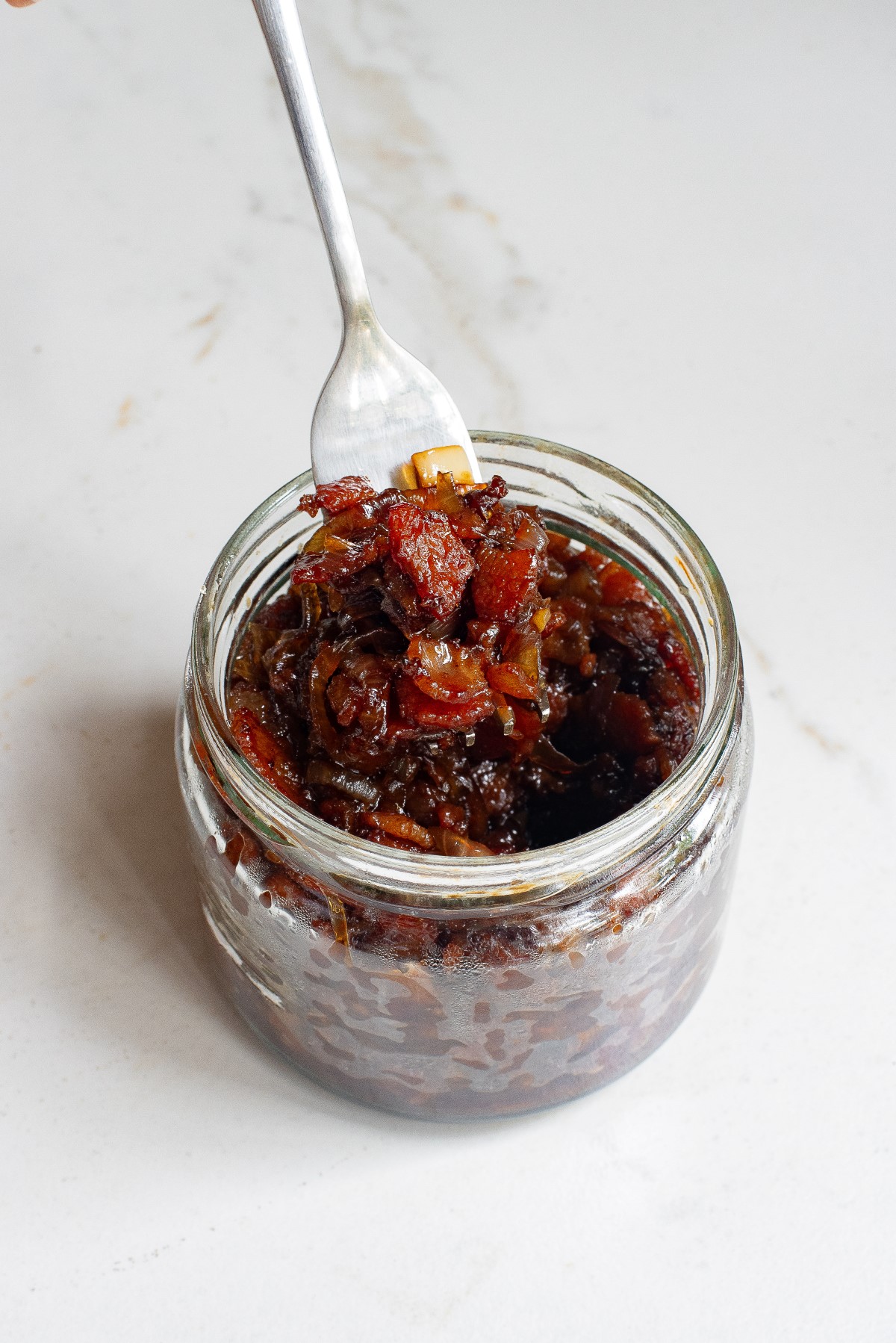 Onion Bacon Jam in a small glass jar with a fork lifting some of the jam out of the jar.