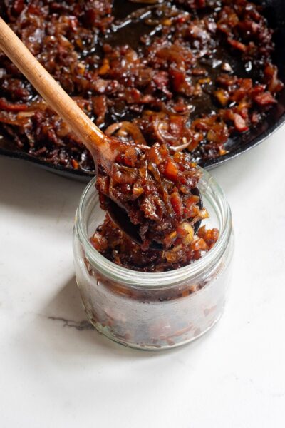 Onion bacon jam being spooned into a small glass jar from the skillet with a wooden spoon.