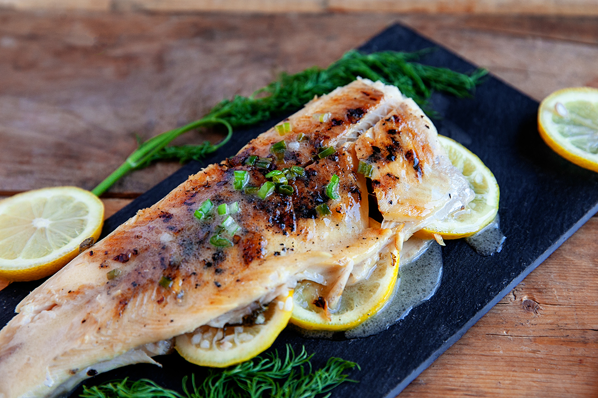 Grilled rainbow trout on a black platter covered in a lemon garlic butter sauce.