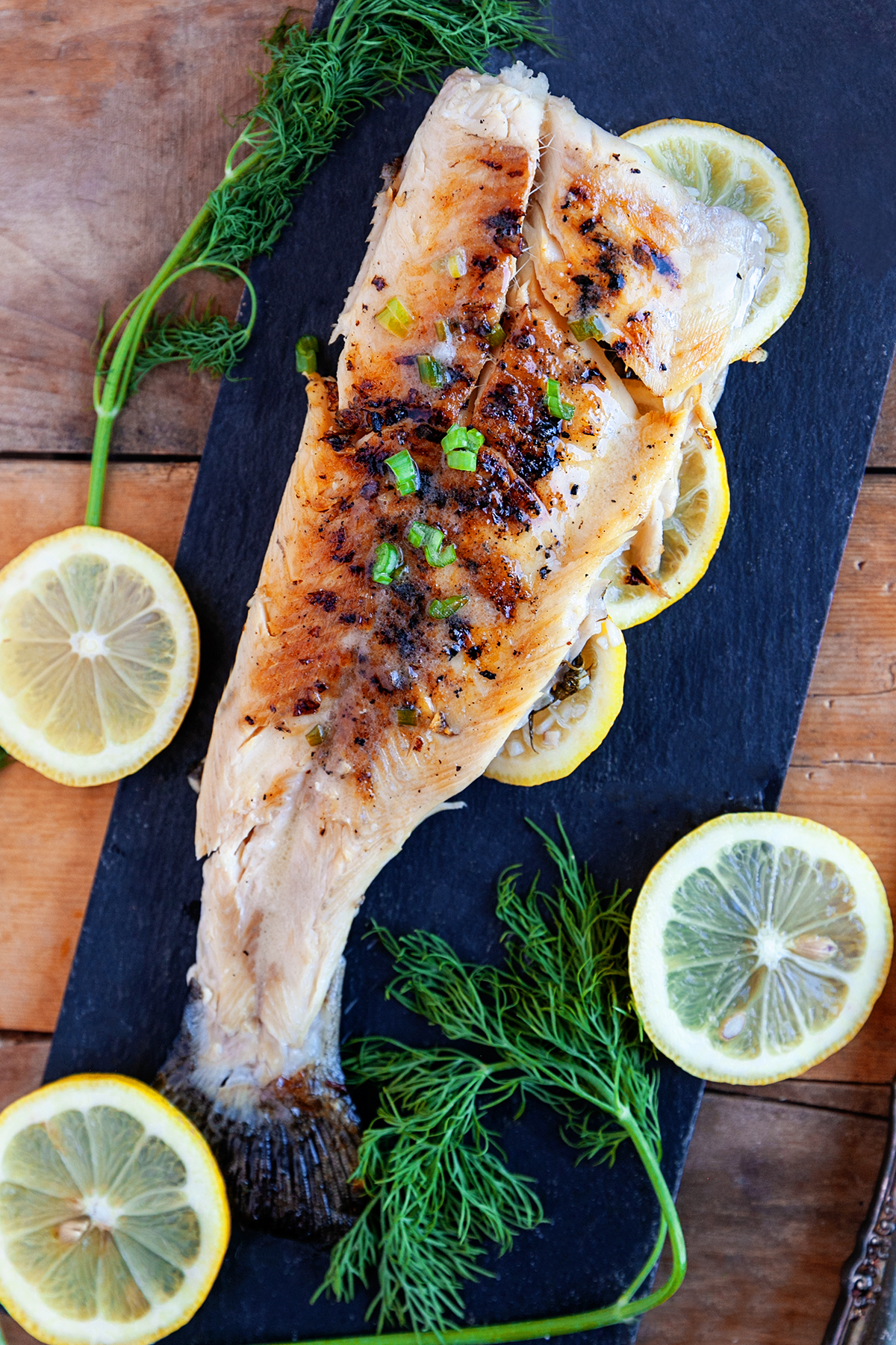 Whole grilled rainbow trout on a black serving board with lemon slices and fresh dill.