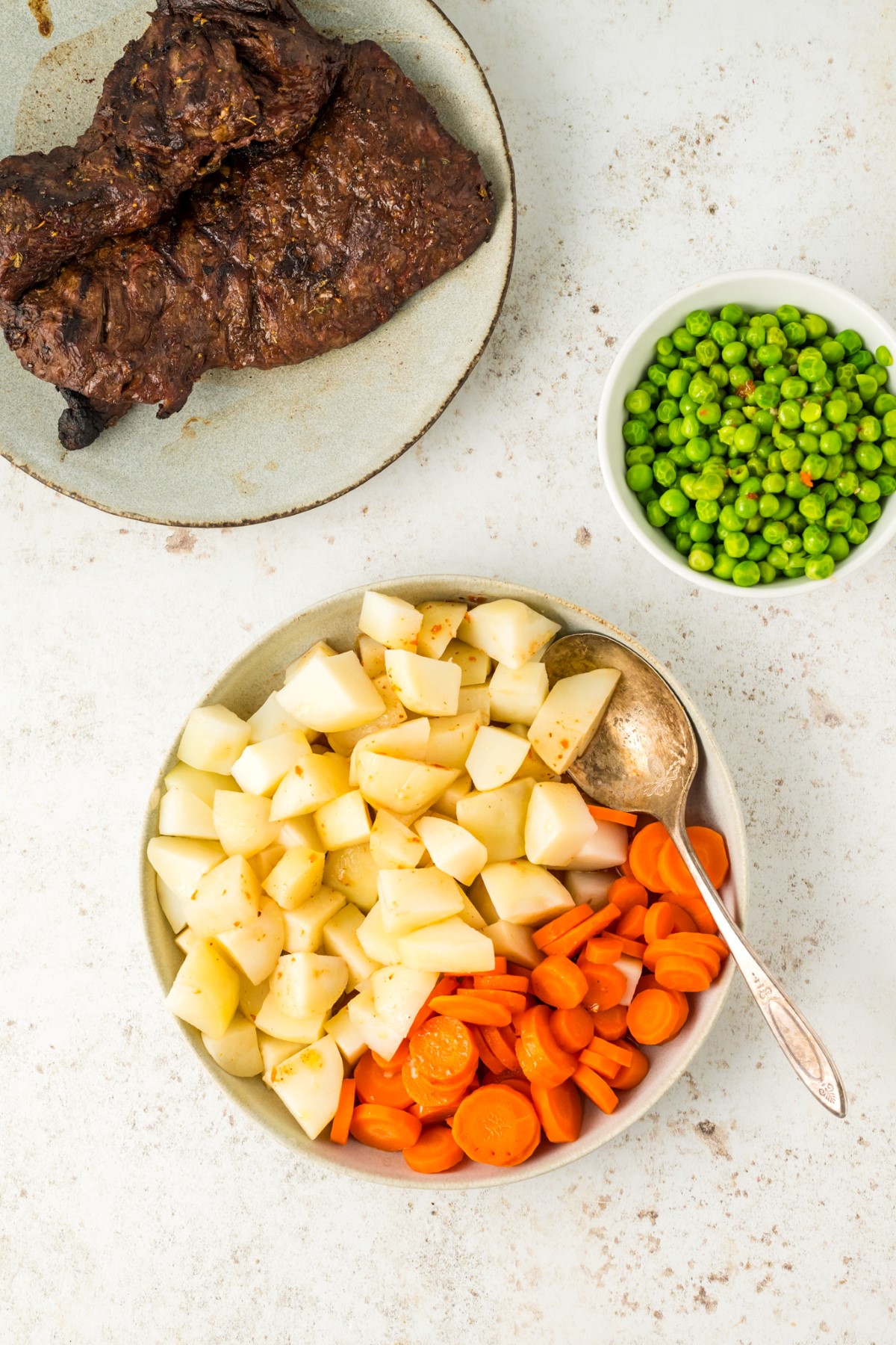 Grilled flank streak, cooked peas, carrots, and potatoes in separate bowls.